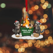 Christmas ornament with two raccoons around a fire, featuring the text 'Feral and Festive', against a blurred festive background.
