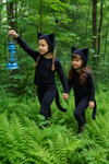 Two children in black cat costumes walking through a forest.